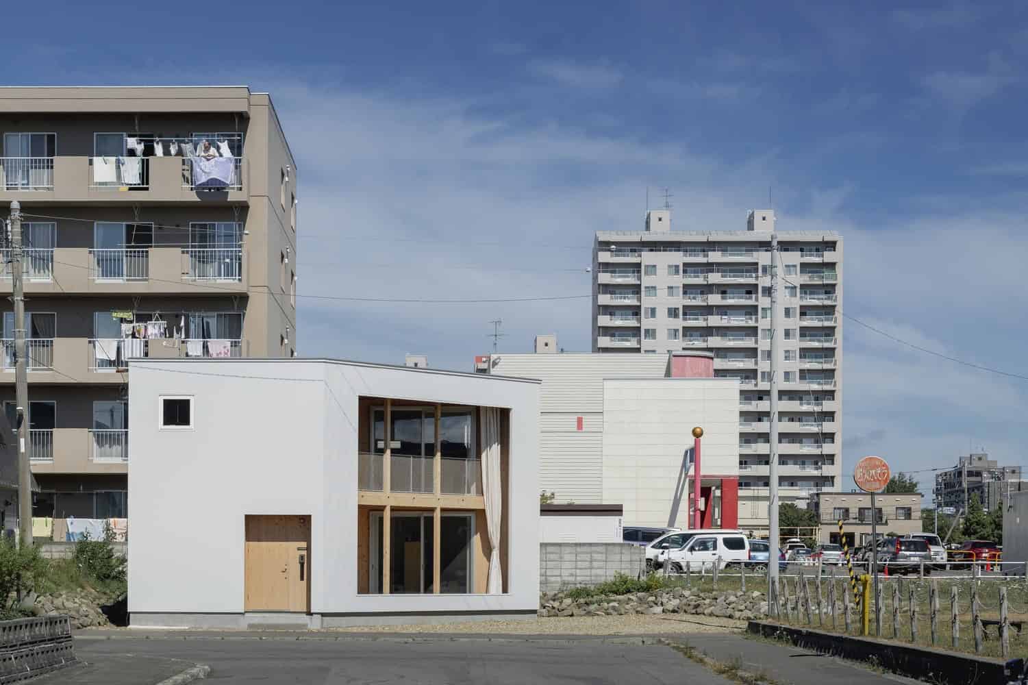 Modern minimalist tiny house with white exterior and large windows, situated in an urban area with high-rise apartment buildings in the background.