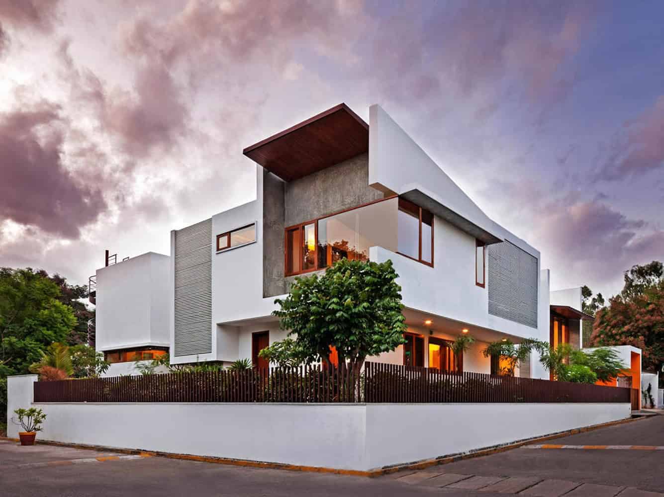 Modern minimalist white house with contemporary architectural design, large windows, and clean lines, surrounded by lush greenery and a stylish fence, under a dramatic evening sky.