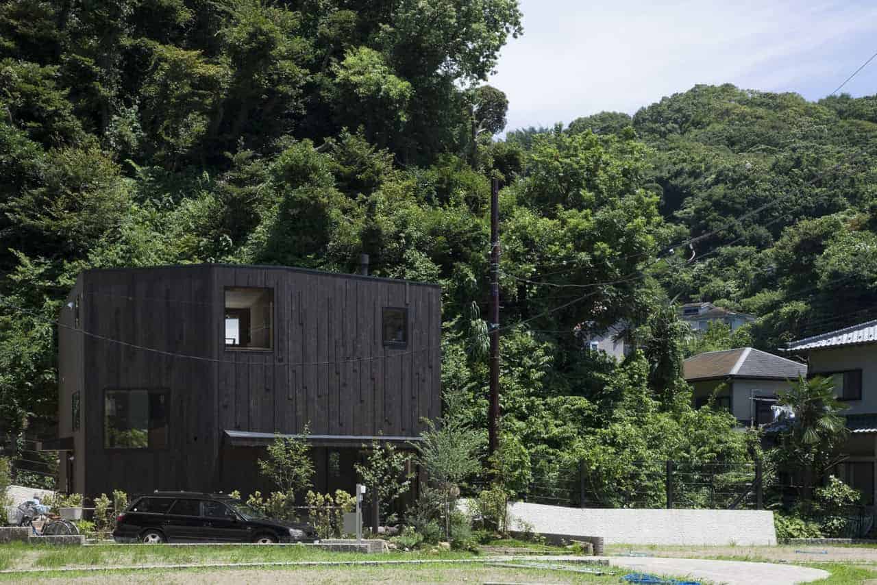 Modern black wooden residential house with unique geometric design, surrounded by lush green trees in a hilly landscape.
