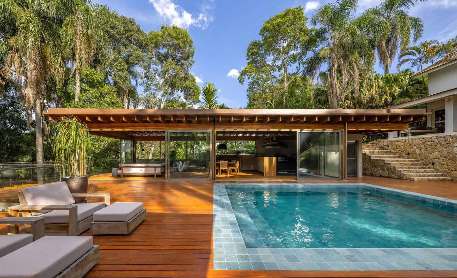 Contemporary outdoor pool area with modern wooden deck, glass sliding doors, lush tropical trees, and cozy lounge furniture, perfect for stylish backyard designs.