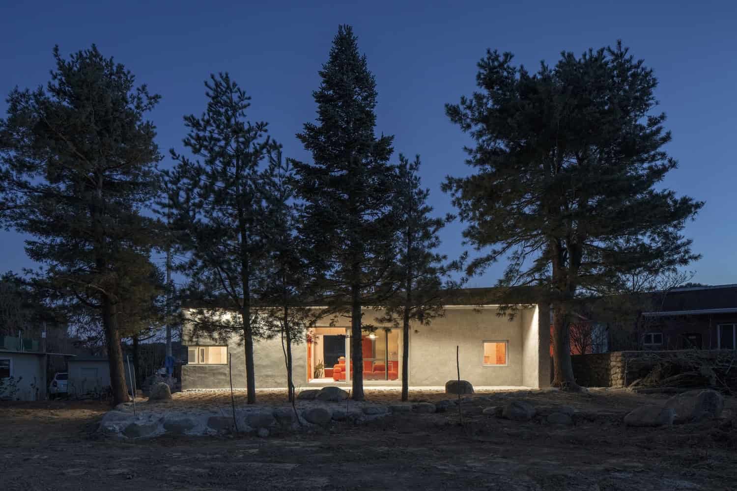 Modern minimalist house surrounded by tall pine trees, contemporary architecture with clean lines, large glass windows, and outdoor lighting in a serene natural setting.