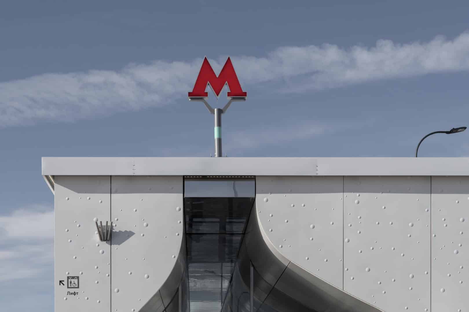 Elevator and metro station entrance with modern architectural design, featuring a sleek white exterior and a red metro symbol on top, against a blue sky background.