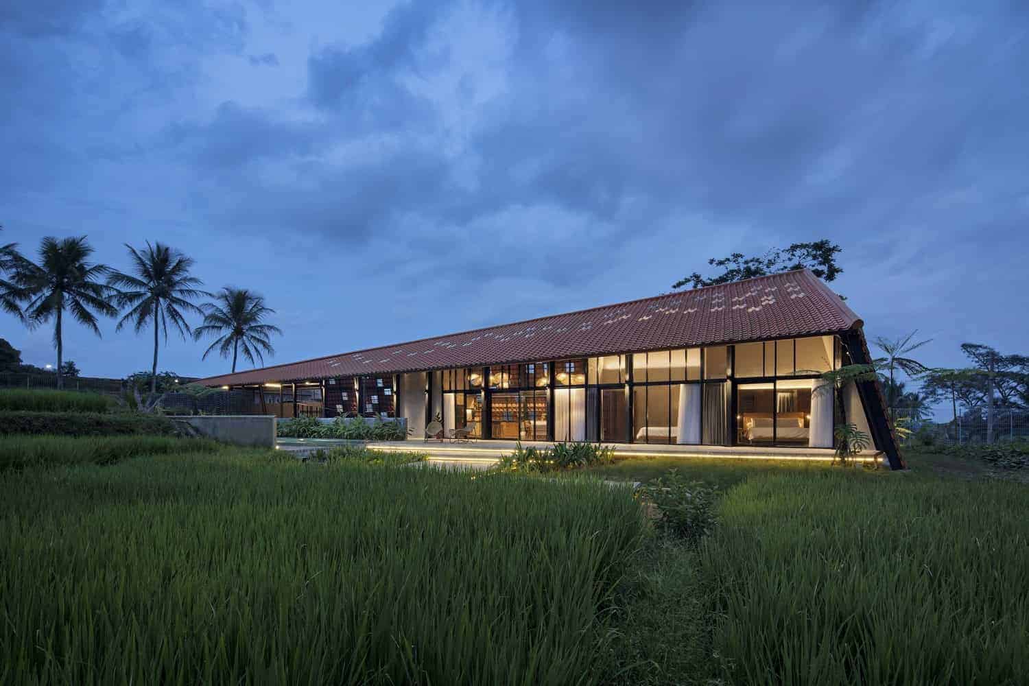 Modern farmhouse with large glass windows and a sloped terra cotta roof set amidst lush greenery and palm trees at dusk.