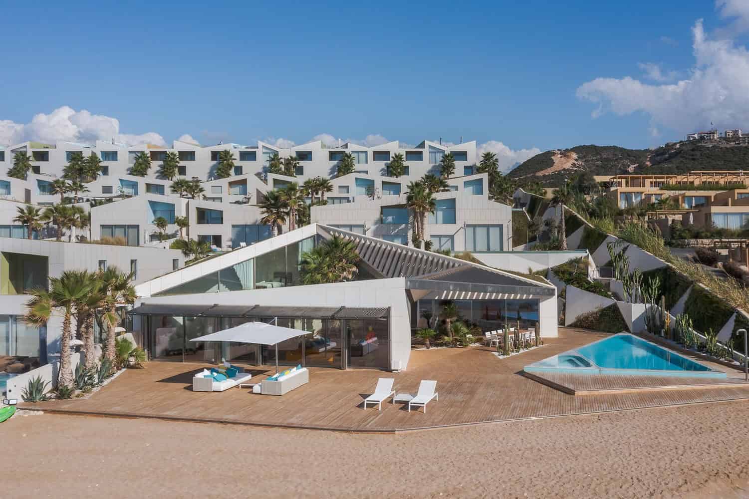 Modern luxury beachfront resort with contemporary white architecture, tropical palm trees, outdoor pool, patio lounge area, and hillside villas in a scenic coastal setting.
