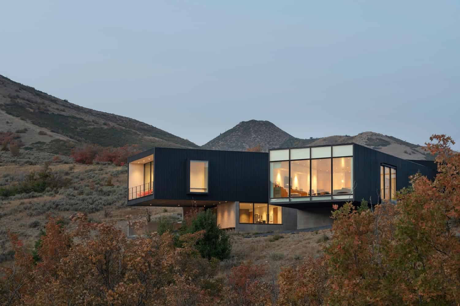 Sleek modern hillside house with large glass windows and contemporary architecture nestled among autumn trees and mountains.
