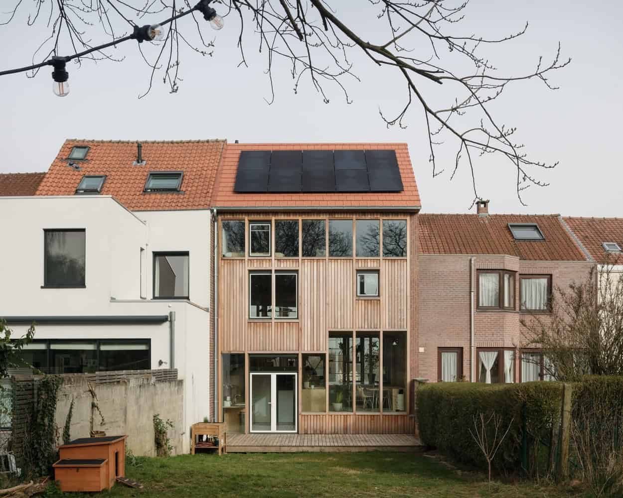Modern multi-story house with solar panels on red tile roof, wooden facade, large windows, and outdoor garden space, showcasing contemporary architecture and sustainable design.