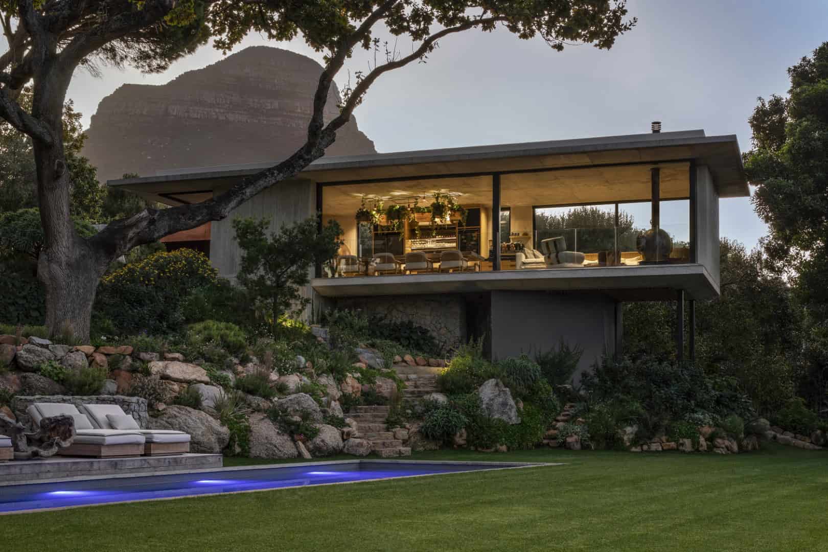 Modern glass architecture home with a sleek design overlooking a lush landscaped garden and pool, surrounded by natural scenery and a mountain backdrop.