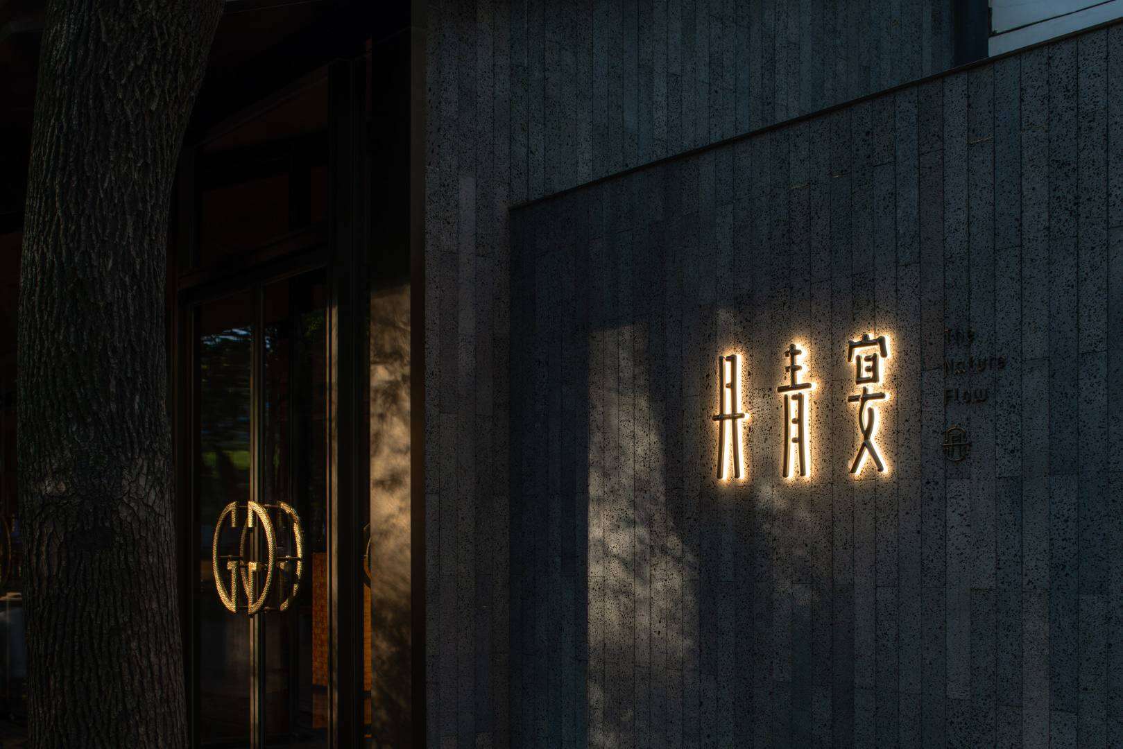 Contemporary building entrance with illuminated Asian-inspired characters and modern architectural design, featuring textured concrete walls and sleek glass doors.