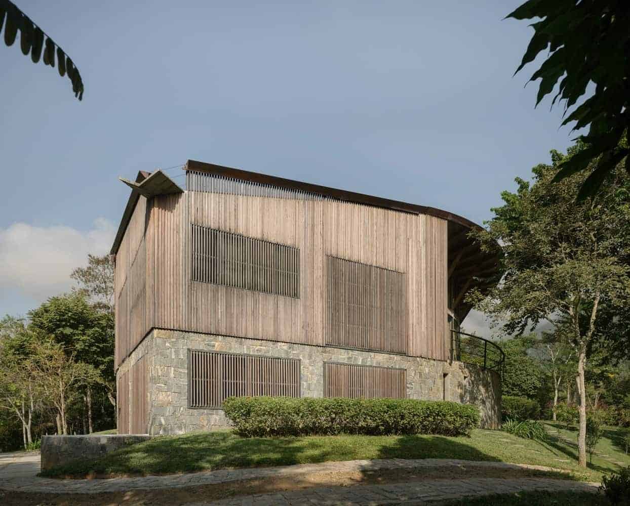 Modern wooden house with stone foundation, featuring horizontal slats and large windows, surrounded by lush greenery and trees, showcasing contemporary architecture and eco-friendly design.