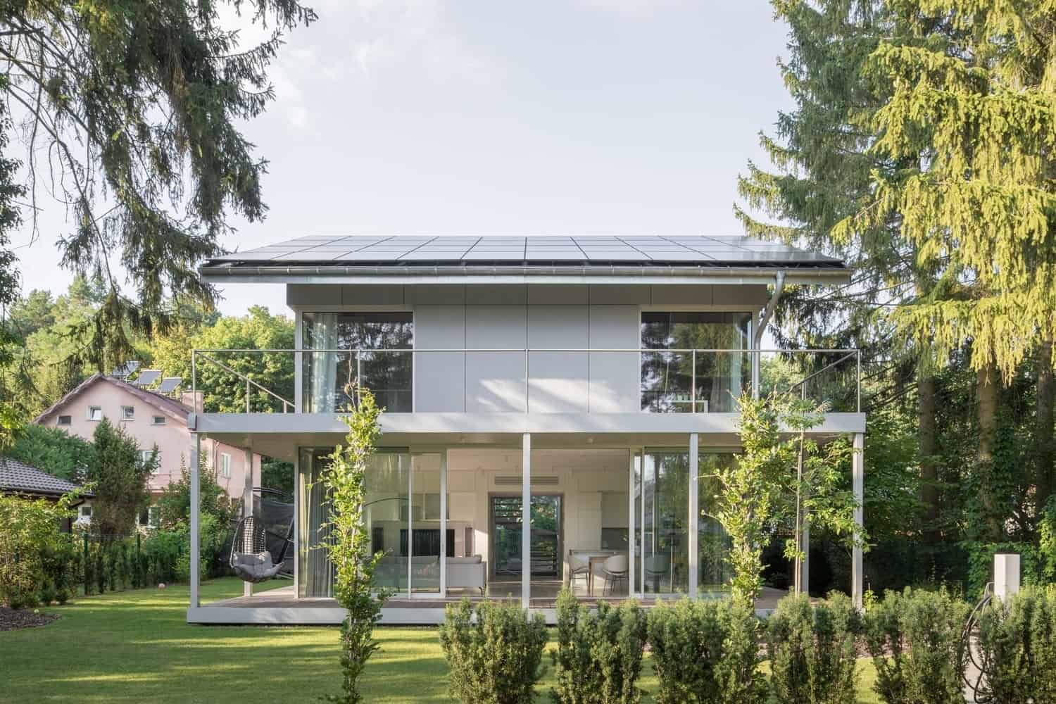 Modern glass house with sustainable solar panels, surrounded by lush greenery and trees, showcasing innovative and eco-friendly architecture.
