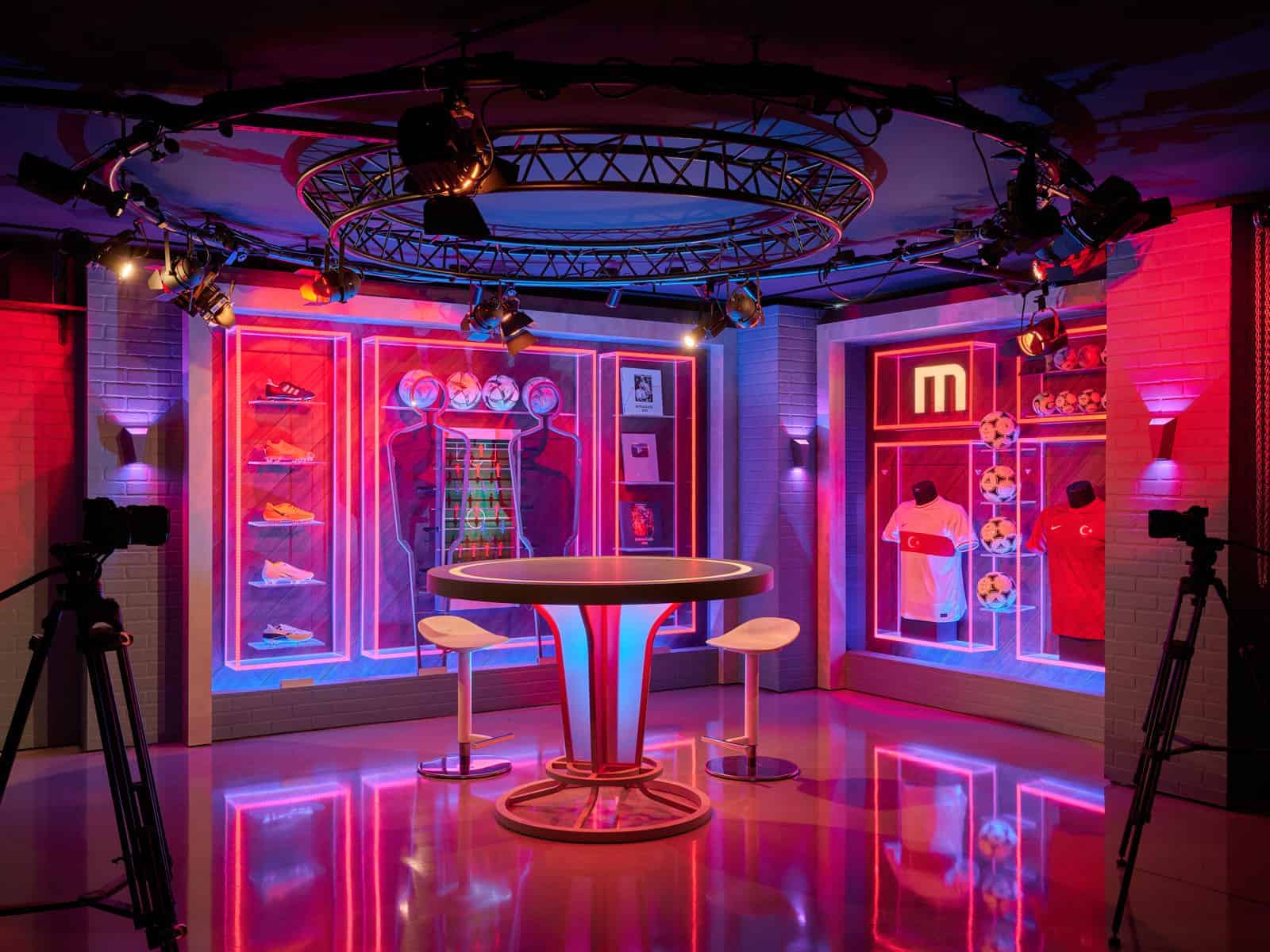 Vibrant neon-lit sports merchandise store with illuminated displays of sneakers, jerseys, and soccer balls, featuring modern interior design and professional camera setup.
