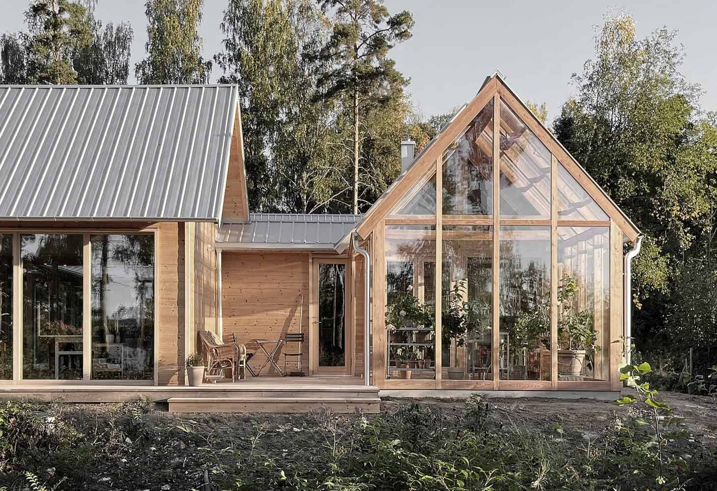 Modern wooden house with greenhouse extension, featuring large glass windows and metal roof, surrounded by nature, showcasing innovative architecture and eco-friendly design.