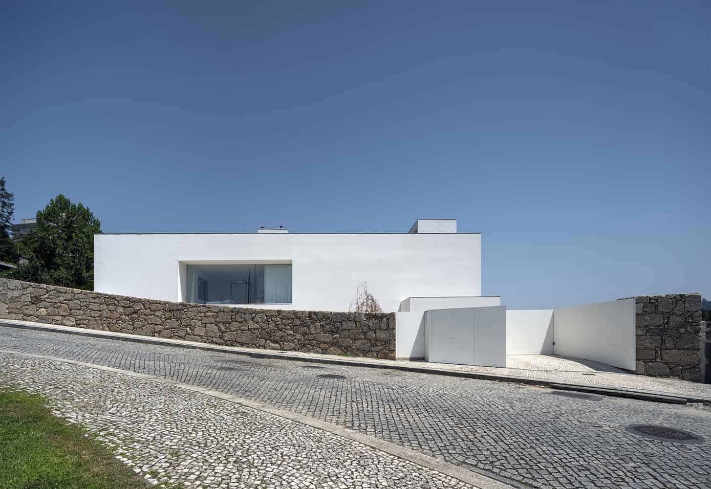 Modern minimalist white house with clean lines and large glass windows, situated on a sloped cobblestone street with stone walls, showcasing contemporary architecture and innovative design elements.