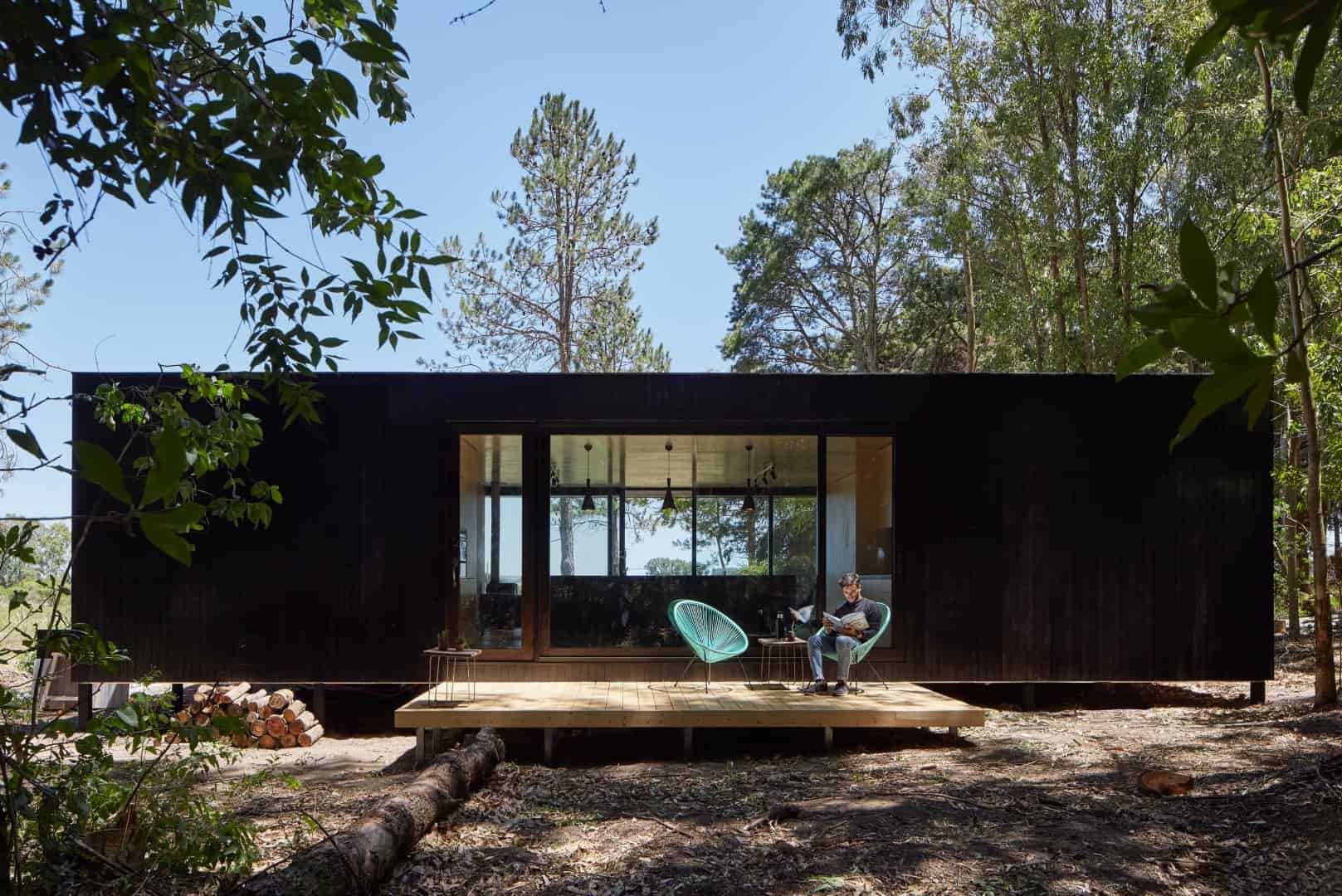 Contemporary black cabin with large glass windows surrounded by lush green trees and forest, set on a wooden deck with minimalist outdoor furniture.