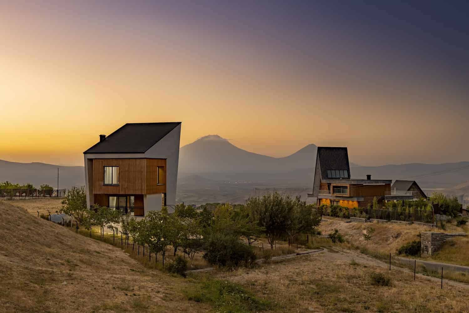 Contemporary modern houses with unique architectural design and sleek wooden facades against a sunset sky with mountains in the background.
