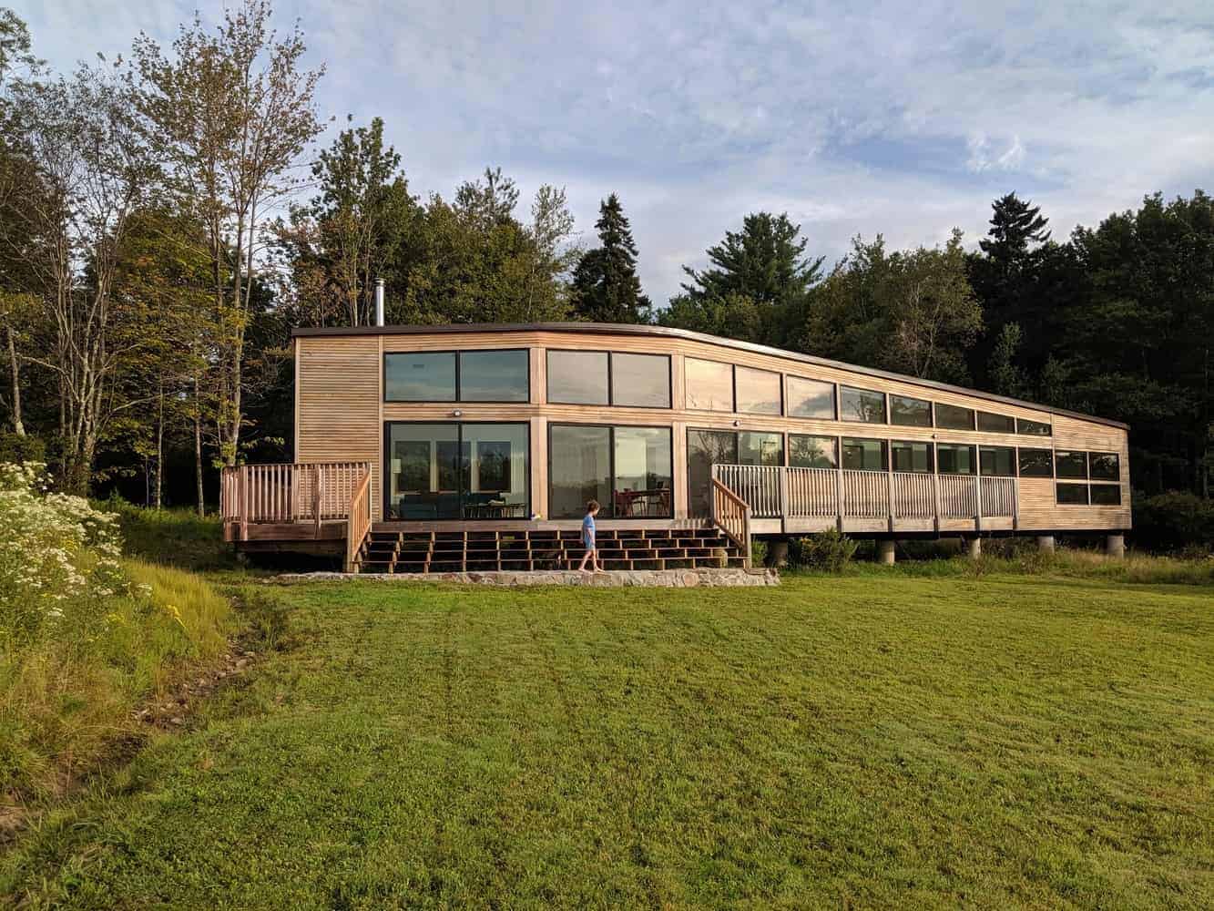 Modern wooden house with large glass windows and surrounding deck on grassy terrain, nestled among trees, showcasing contemporary architecture and eco-friendly design elements.