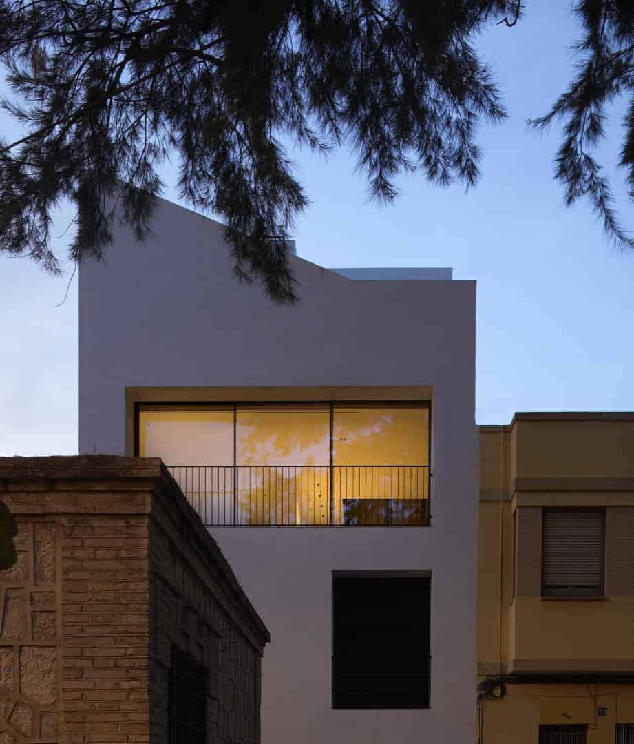 Modern minimalist white residential building with large glass windows and black metal balcony, surrounded by trees, showcasing contemporary architecture and urban design.