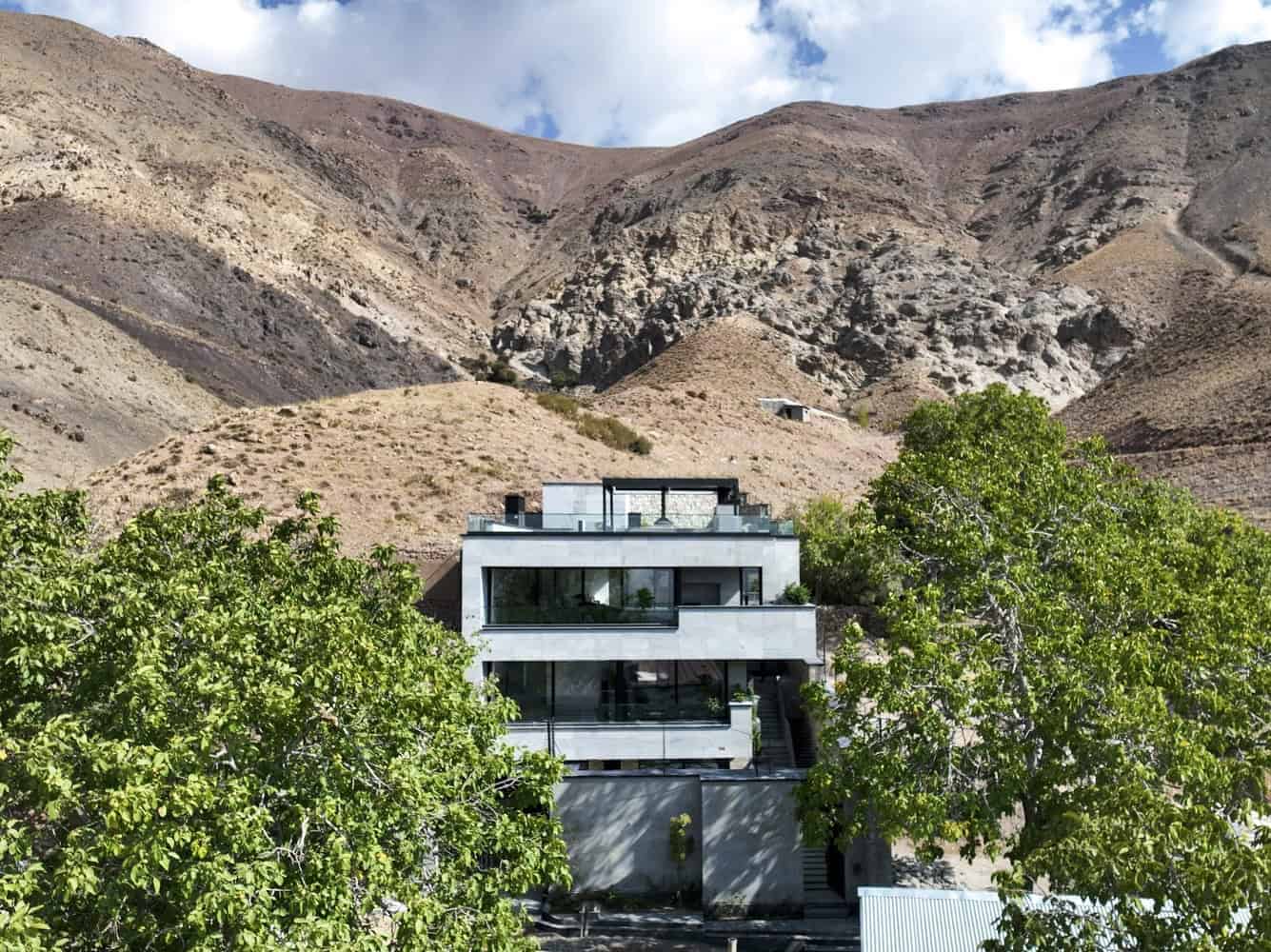 Modern minimalist house with large glass windows and terraces, nestled amidst lush green trees in a mountainous landscape, showcasing contemporary architecture and scenic natural surroundings.