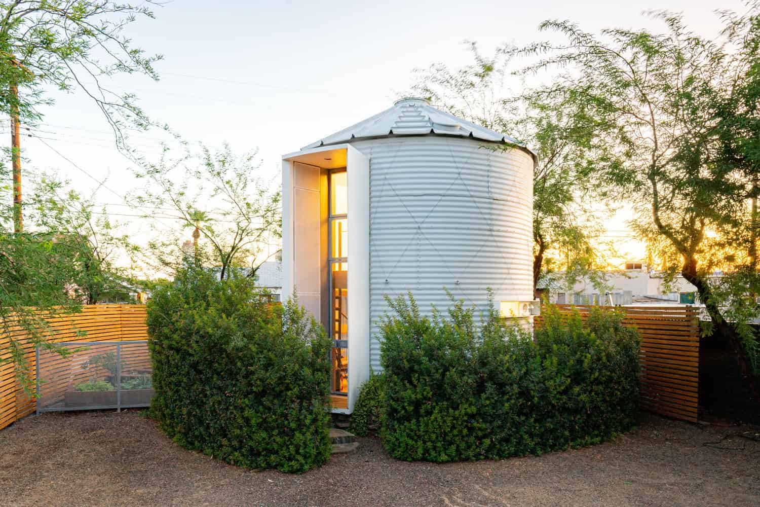 Modern tiny house with a unique cylindrical design, surrounded by greenery and wooden fencing, showcasing innovative architecture and sustainable living concepts.