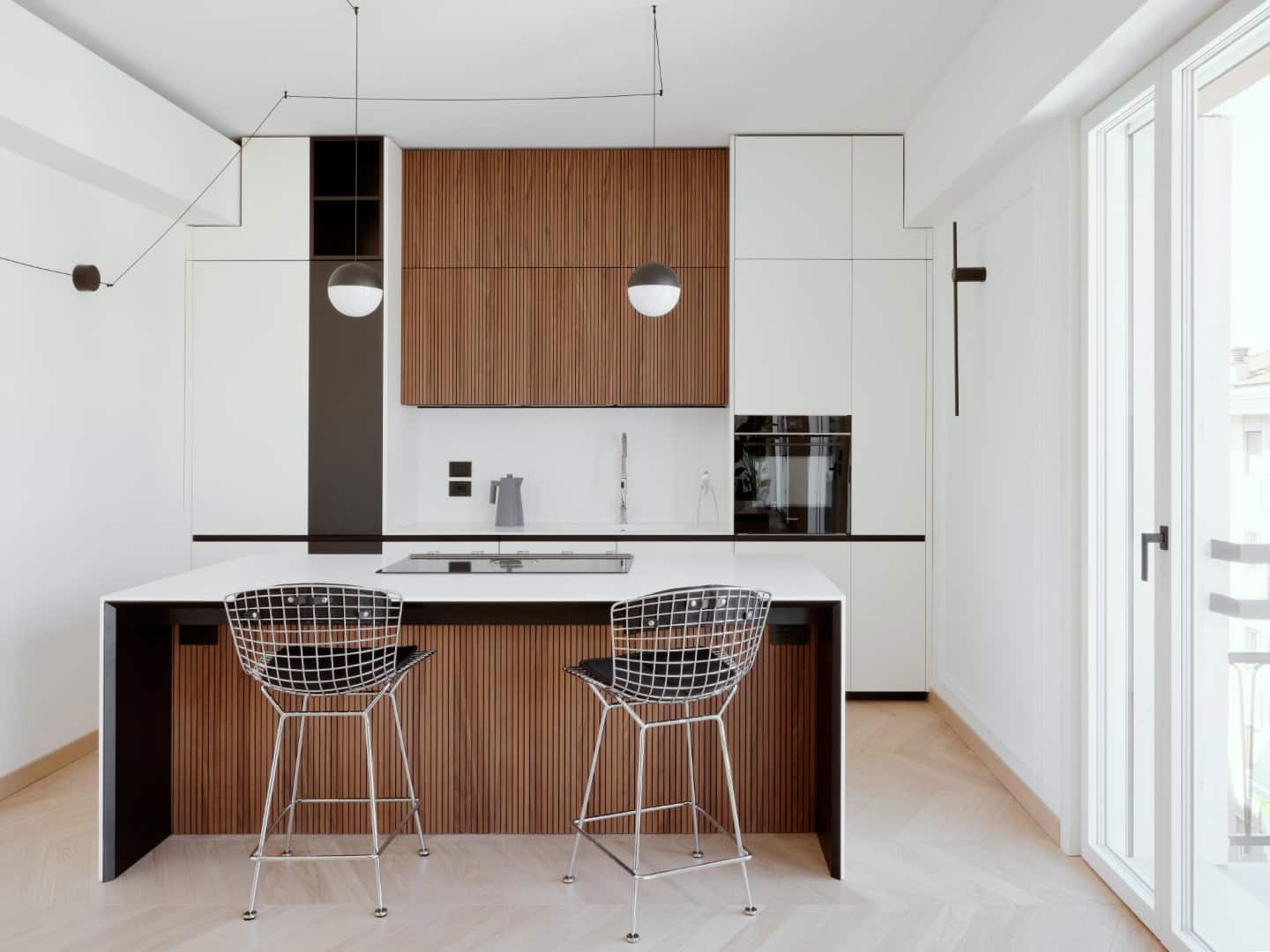 Sleek modern kitchen with minimalist design, wooden cabinet accents, and contemporary pendant lighting. Bright and airy space with clean lines and stylish black wire chairs.