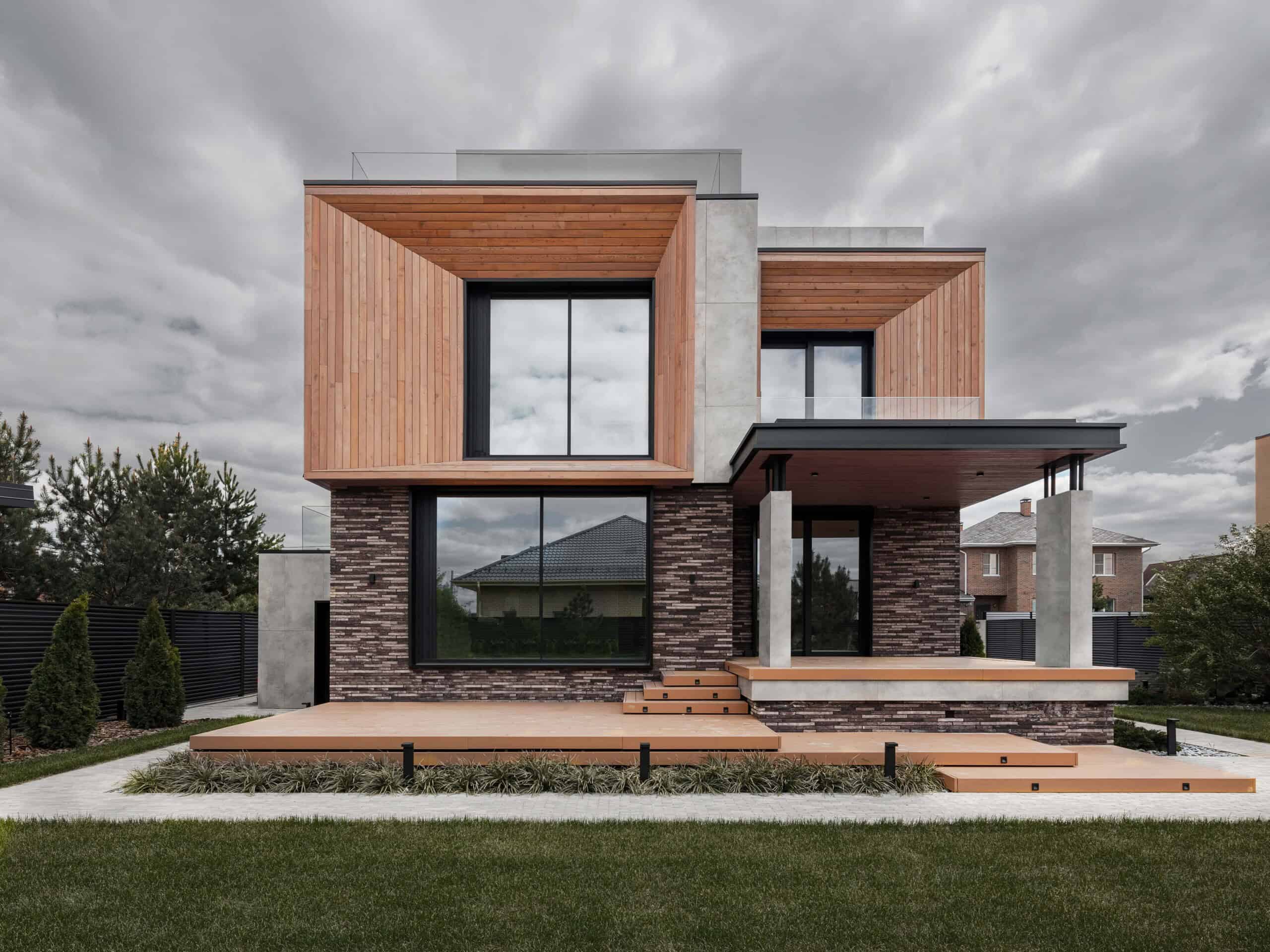 Contemporary modern house with wood and stone exterior, large windows, and a minimalist design, surrounded by a landscaped yard under a cloudy sky.