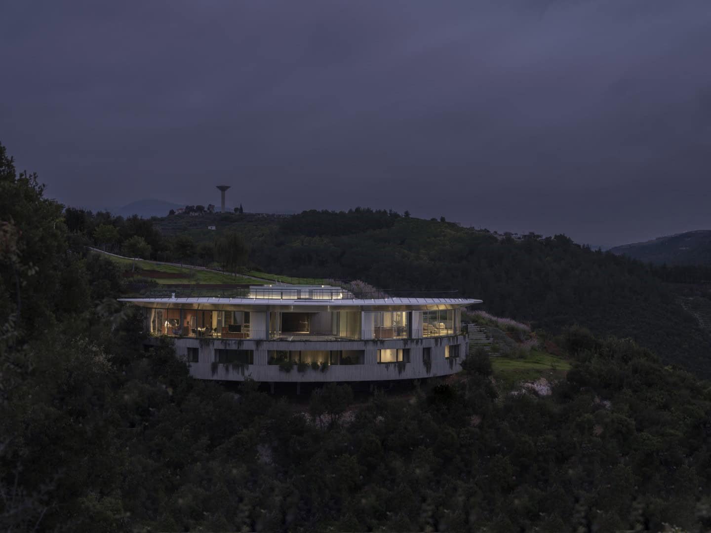 Modern hillside luxury house with floor-to-ceiling windows and sleek architectural design at dusk, surrounded by lush greenery and scenic mountain views.