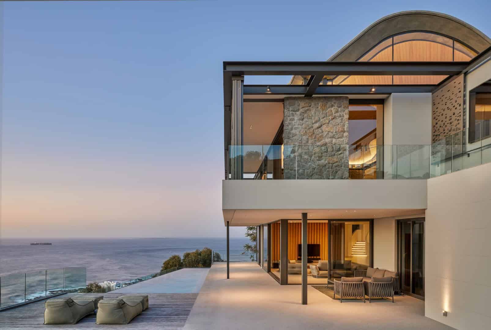 Modern luxury house with panoramic ocean views, contemporary architecture, glass balcony, stone accents, and spacious outdoor terrace at sunset.