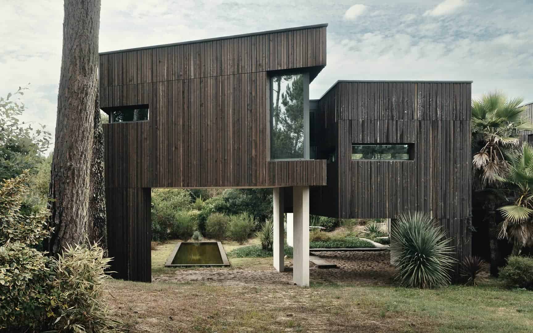 Sleek modern house with dark wooden exterior, geometric architecture, large windows, surrounded by lush greenery and landscaped garden, showcasing contemporary minimalist design.