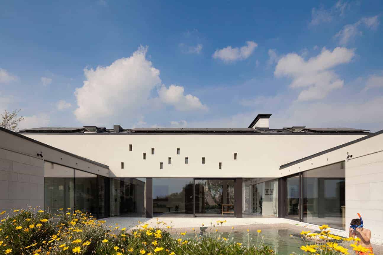 Modern minimalist house with clean lines, large glass windows, and contemporary architecture, set against a bright blue sky with landscaping including yellow flowers.