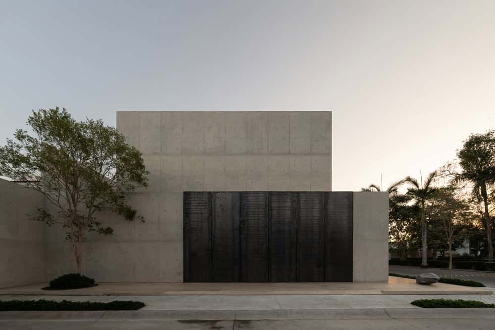 Concrete modern minimalist building with black metal door, surrounded by trees and landscaped greenery, during sunset.