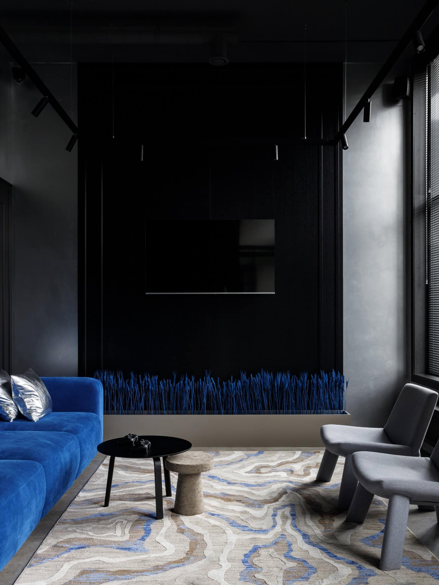 Contemporary modern living room interior with dark walls, vibrant blue velvet sofa, minimalist coffee table, and stylish gray chairs, showcasing sleek modern architecture and design elements.