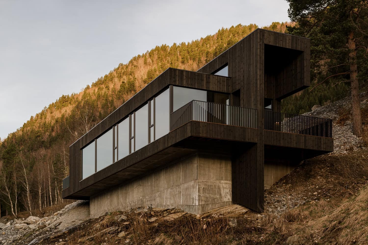 Contemporary black wooden house on stilts with large windows and balcony, situated on a hillside surrounded by trees, showcasing modern architectural design and innovative construction techniques.