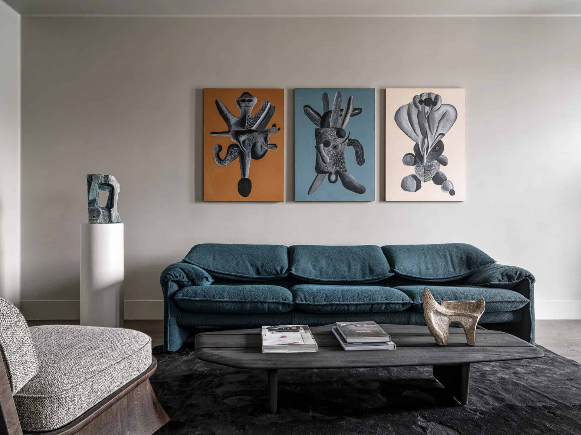 Colorful abstract art paintings hanging above a modern teal sofa in a contemporary living room. Decor includes sculptural pieces and a wooden coffee table on a dark rug, combining artistic interior design elements.