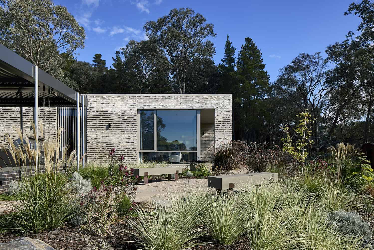 Contemporary modern house with minimalist architecture, flat roof, and white brick exterior, surrounded by desert landscaping with ornamental grasses and trees under a clear blue sky.