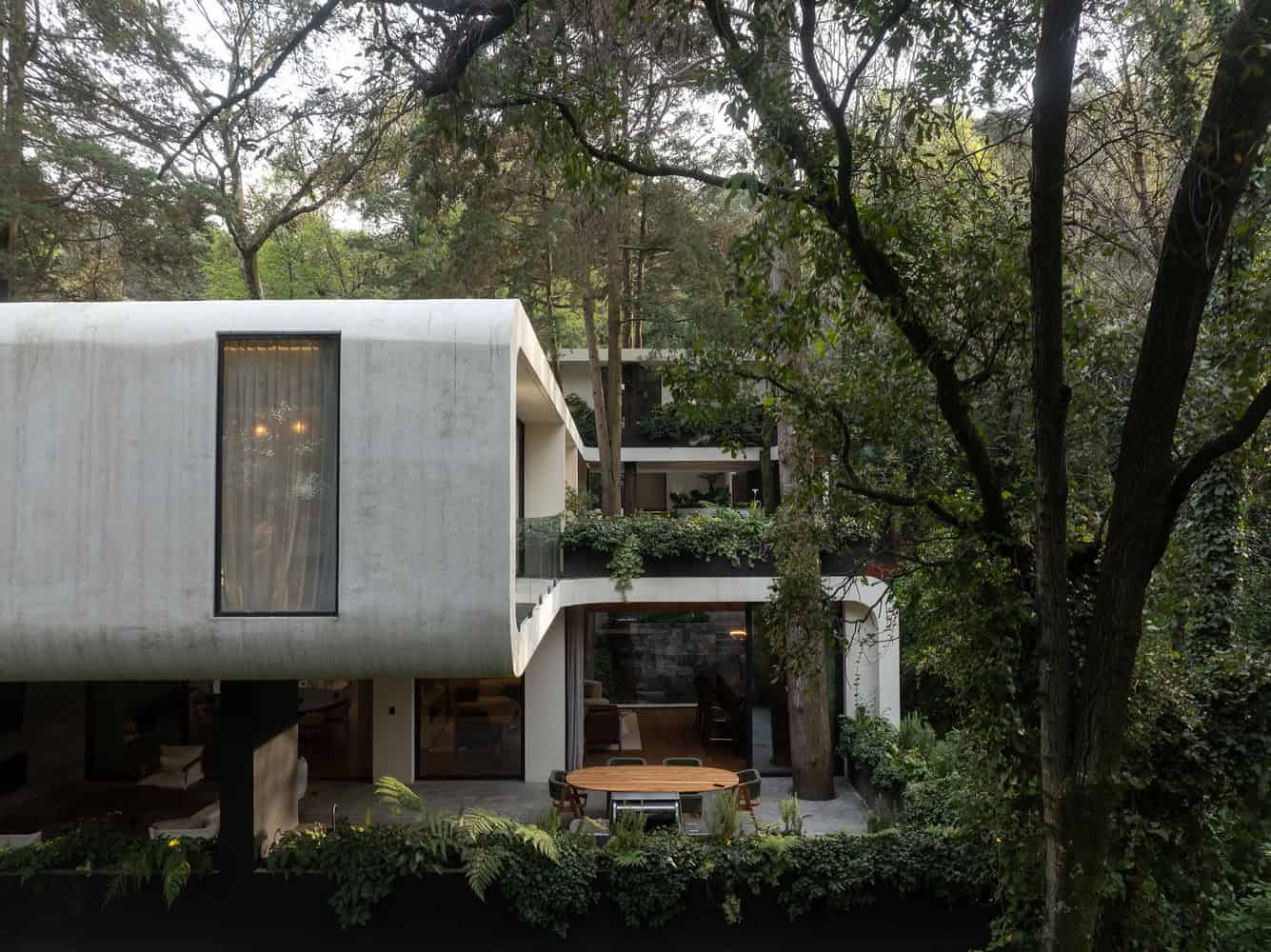 Modern minimalist house with concrete exterior and large glass windows nestled in a lush forest setting, featuring outdoor dining space surrounded by greenery and trees.