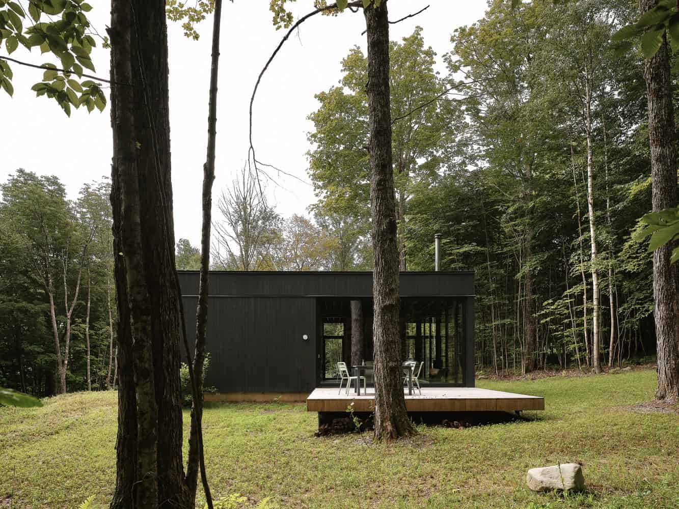 Modern black cabin in tranquil forest setting with wooden deck and outdoor dining area. Surrounded by tall trees, blending minimalist architecture with nature for a peaceful retreat.