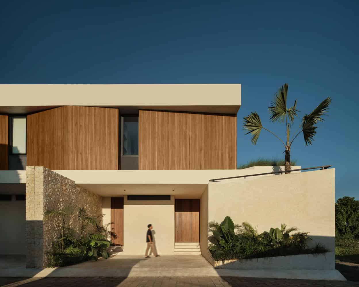 Modern minimalist house with wood paneling, stone accents, and lush landscaping under a clear blue sky.