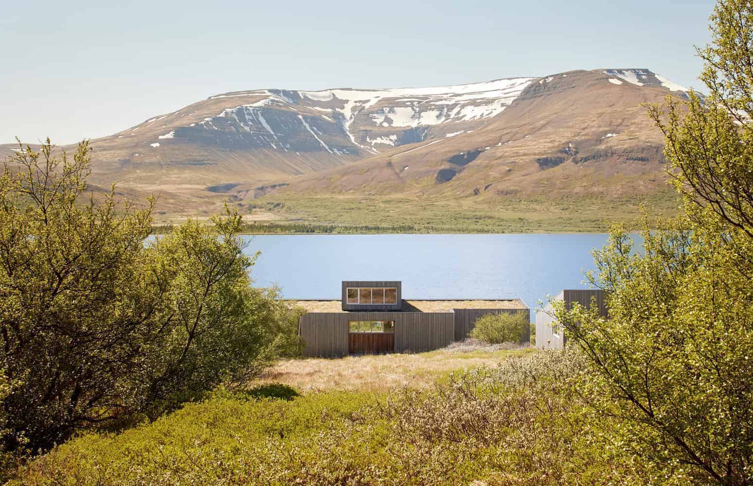 Modern lakeside house with minimalist design, surrounded by lush greenery and overlooking snow-capped mountains and a calm lake, exemplifying contemporary architecture in a natural setting.