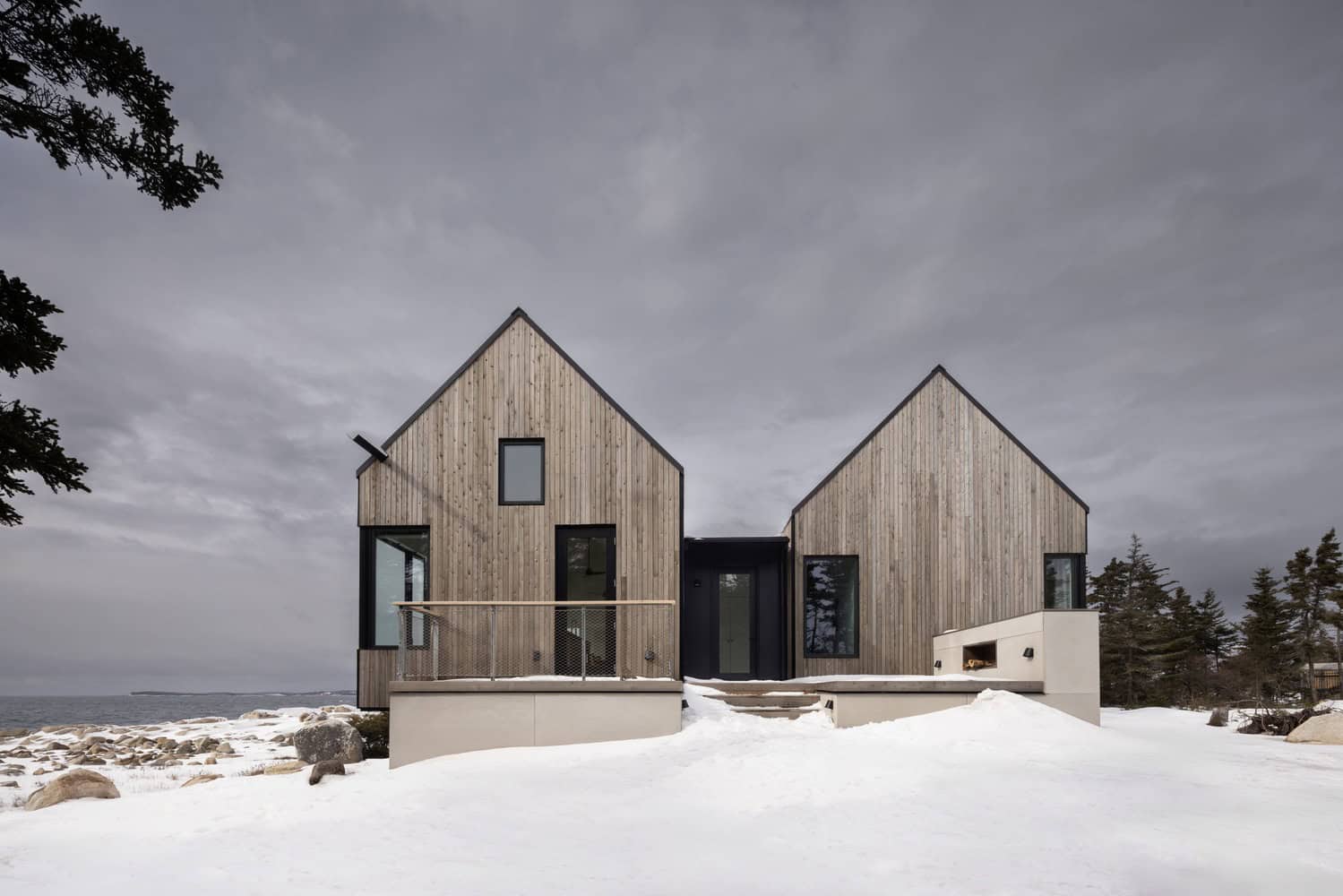 Contemporary wooden house with modern design overlooking a snowy landscape and ocean, featuring minimalist architecture, large windows, and gabled roofs, ideal for winter seaside living.