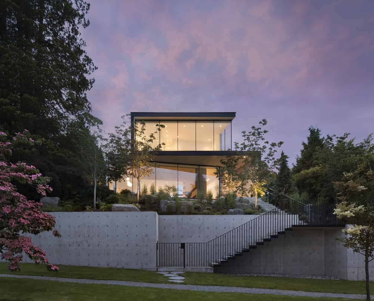 Contemporary modern house with large glass windows, surrounded by landscaped garden and trees, overlooking a colorful sunset sky. Architectural design emphasizes sleek lines, open spaces, and natural integration.