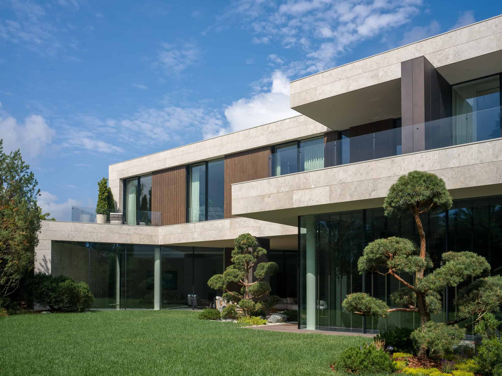 Contemporary villa with large glass windows, lush green lawn, and sculpted trees under a blue sky, showcasing modern architecture and outdoor design.