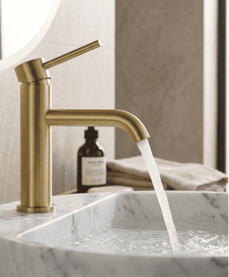 Un rubinetto di ottone con acqua che scorre in un lavandino di marmo bianco; sullo sfondo una bottiglia marrone e asciugamani piegati.