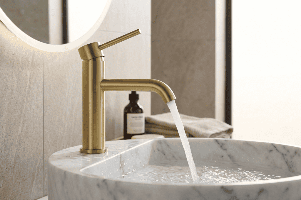 Un moderno rubinetto dorato eroga acqua in un lavandino rotondo di marmo in un bagno con pareti piastrellate beige; sullo sfondo una bottiglia marrone e asciugamani piegati.