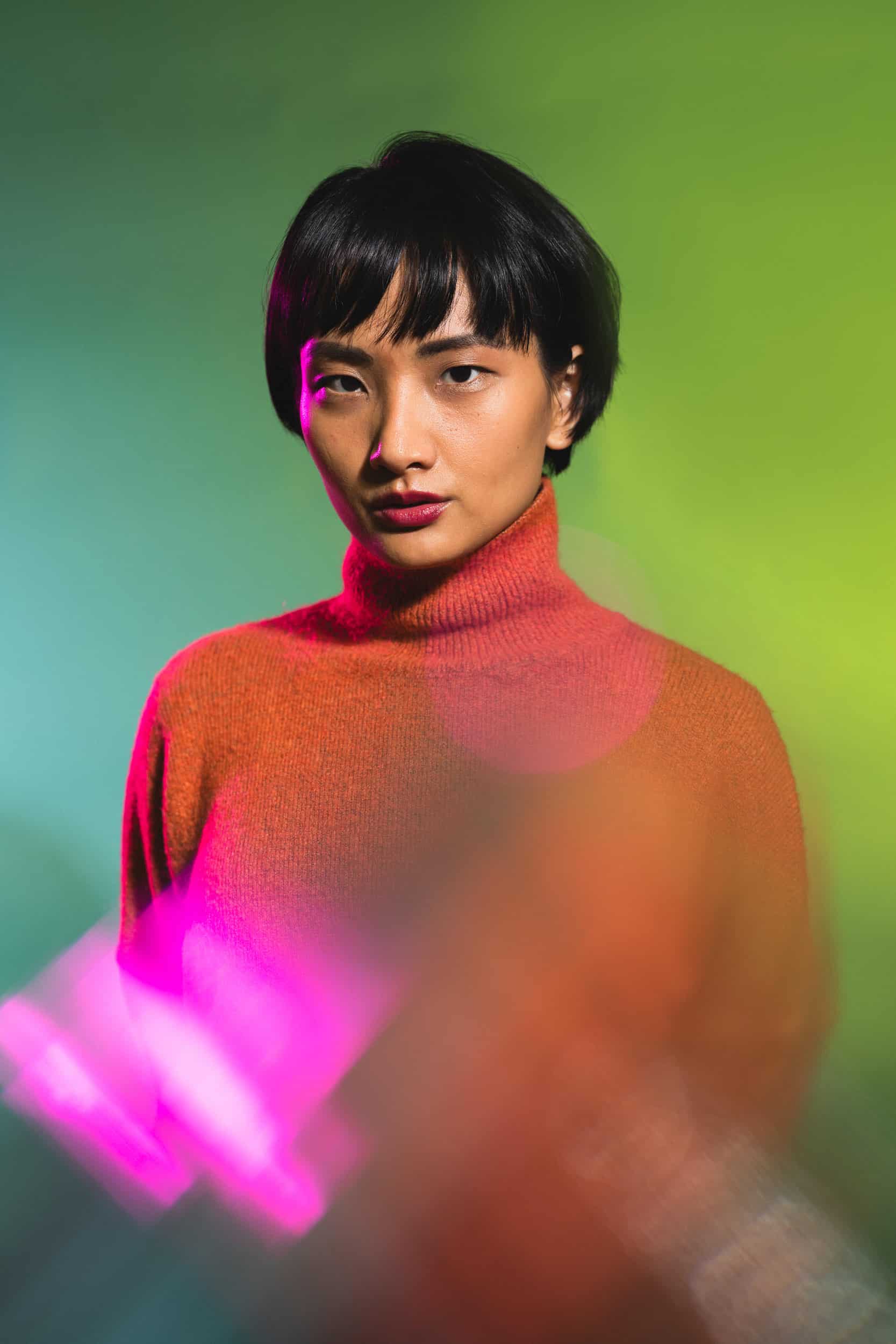colourful-headshot-of-asian-female-in orange-polo-neck-jumper-and-green-background-with-magenta-reflections-in-foreground