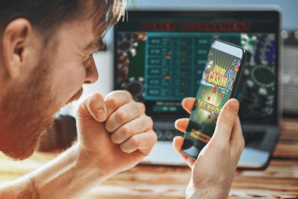 Parental Advice: Mastering Your Child’s Gaming Habits for a Balanced Life 1 A man holds a smartphone displaying an online casino game with the word "JACKPOT," while a poker tournament is visible on a laptop screen in the background.