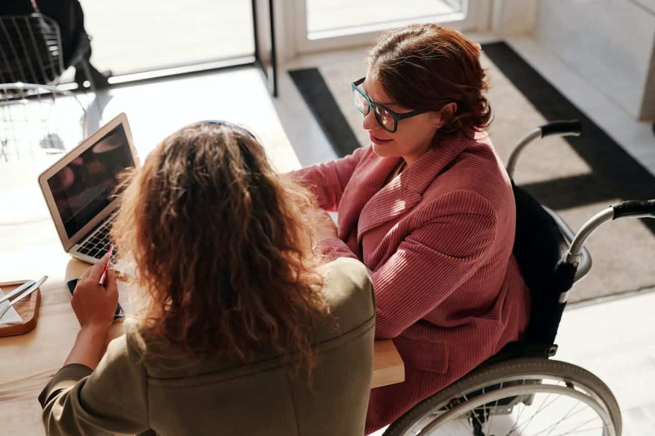 AODA Accesibility for Ontarians with Disabilities Act - two women in front of a laptop. One of them is wearing a red sweater and black framed glasses while seated on a wheelchair. The other woman is back facing with curly hair and wearing an olive green blouse