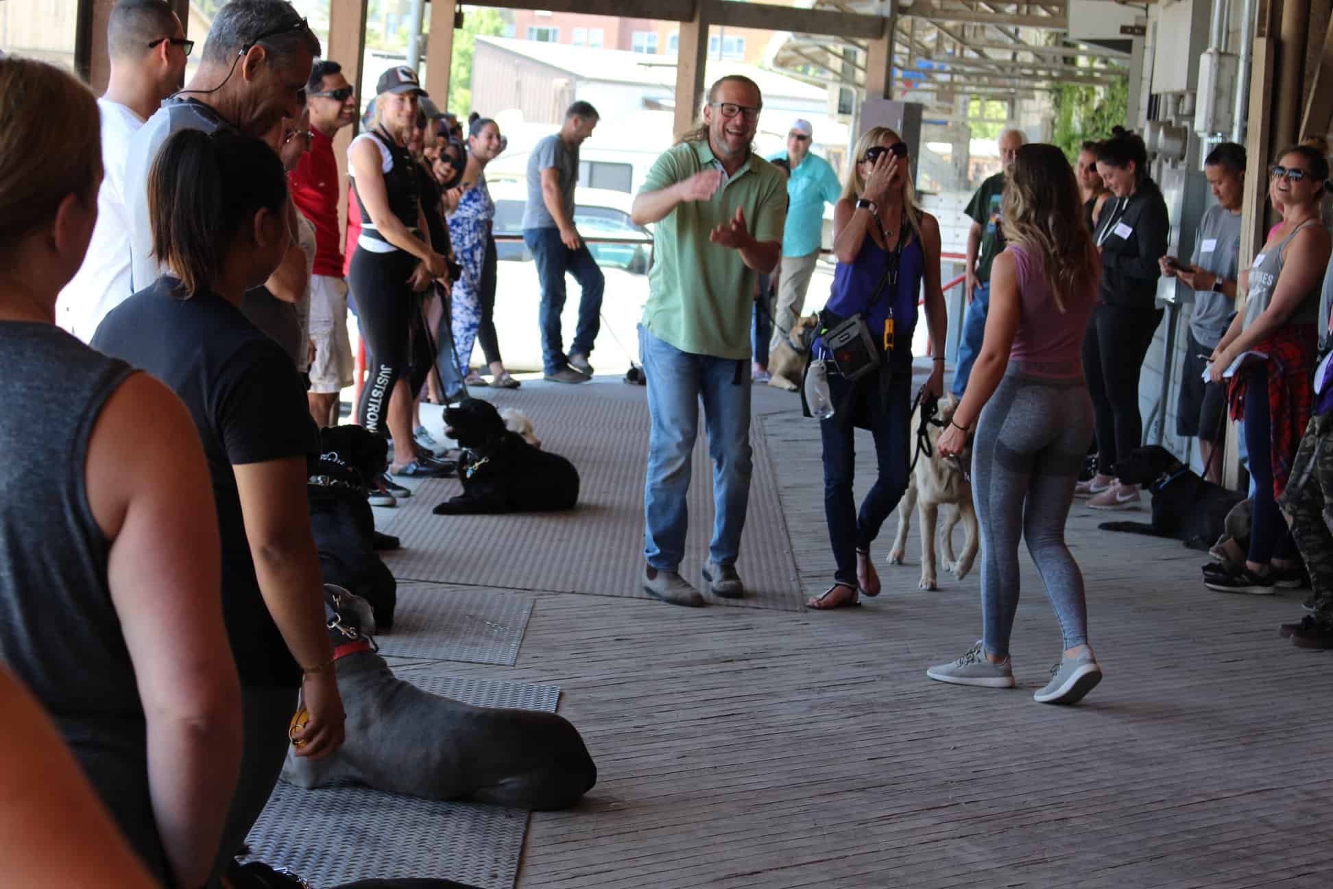 A group of people with dogs gather on a covered wooden deck; one man gestures while others watch or interact with the dogs.