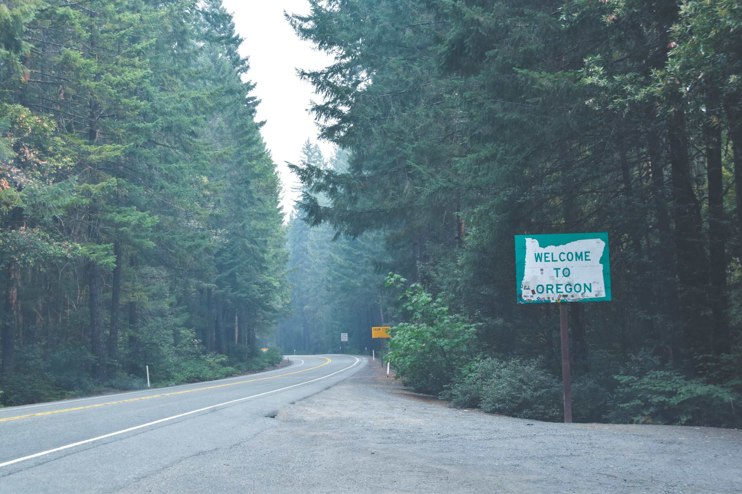 Scenic view of Oregon border with tall pine trees, winding road, and "Welcome to Oregon" sign.