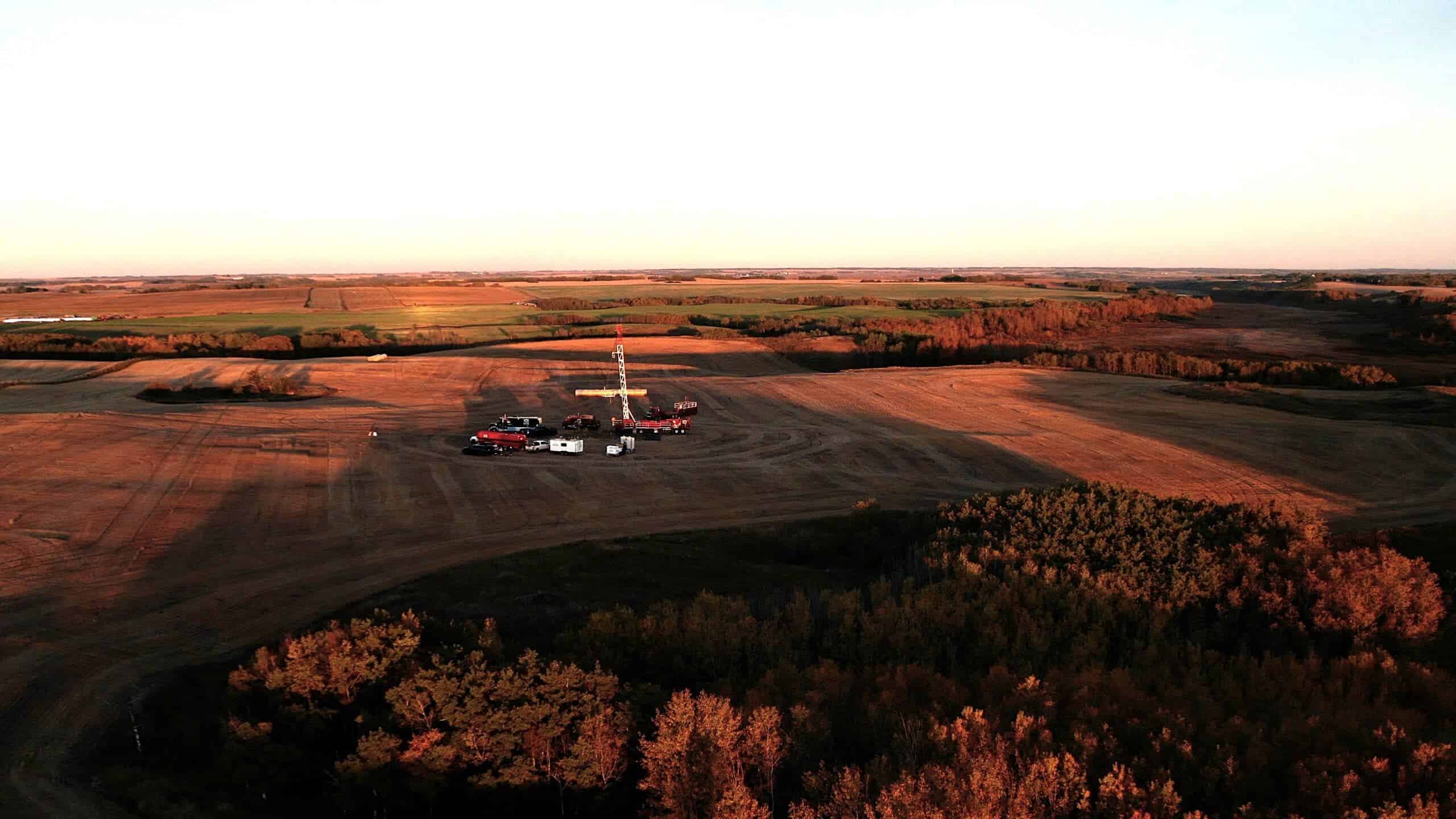Oil drilling rig on farmland with fields and trees during sunset.