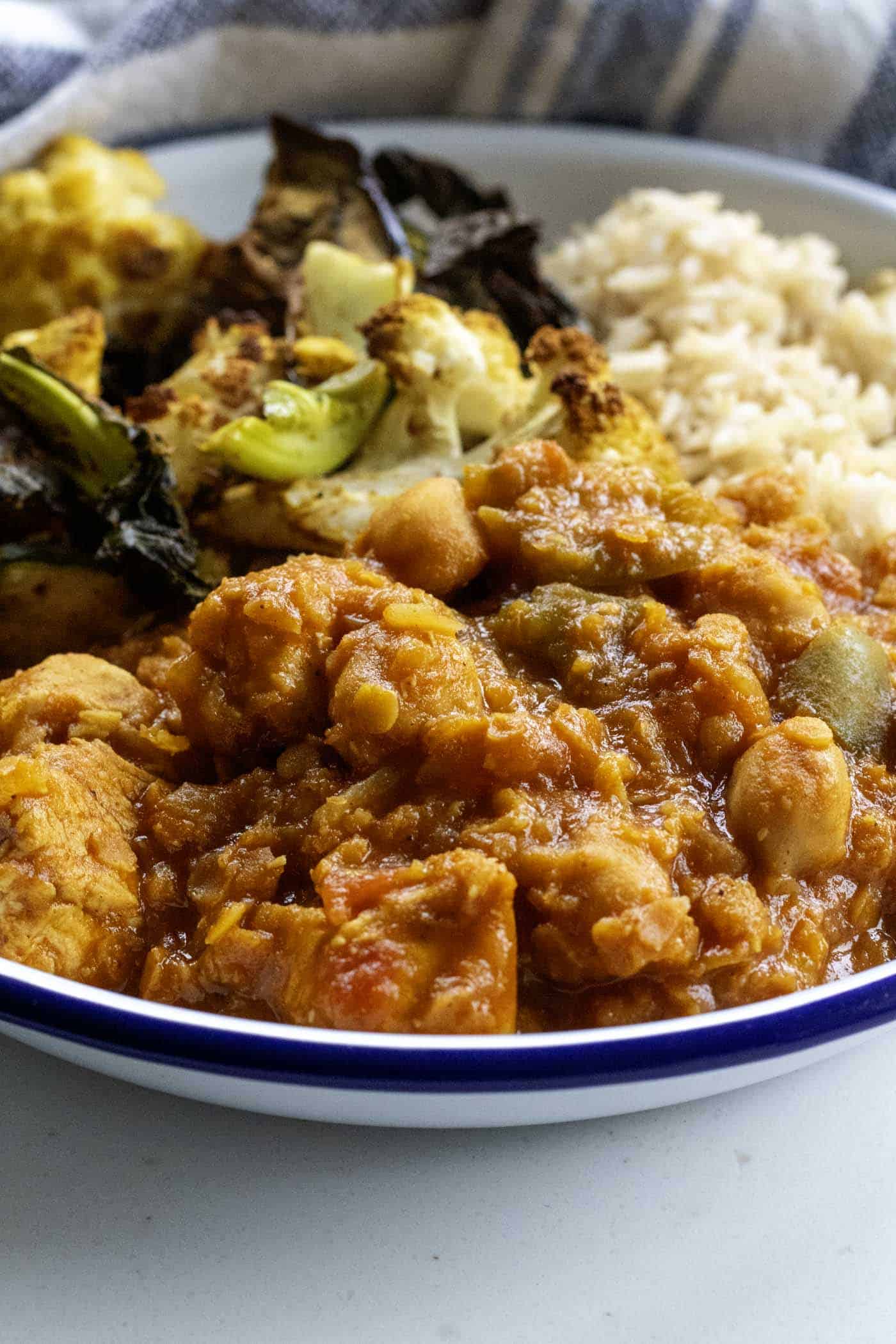 A white bowl filled with rice, vegetables, and a healthy slow cooker chicken curry is placed on a white surface next to a striped cloth.